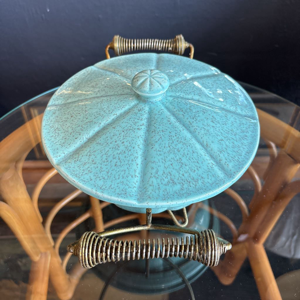 A ceramic turquoise serving bowl and lid in a brass wire caddy with handles on a glass table in front of a black wall.