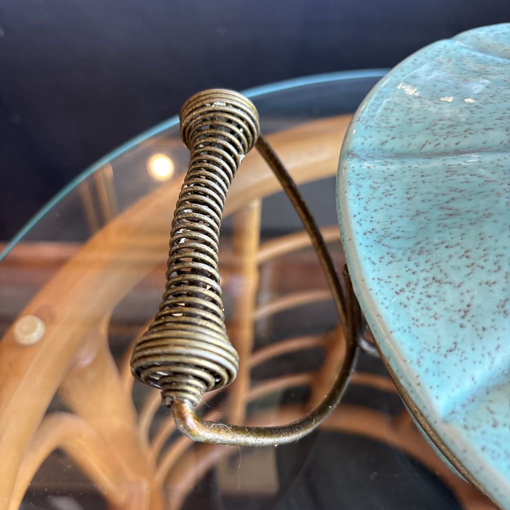 A ceramic turquoise serving bowl and lid in a brass wire caddy with handles on a glass table in front of a black wall.