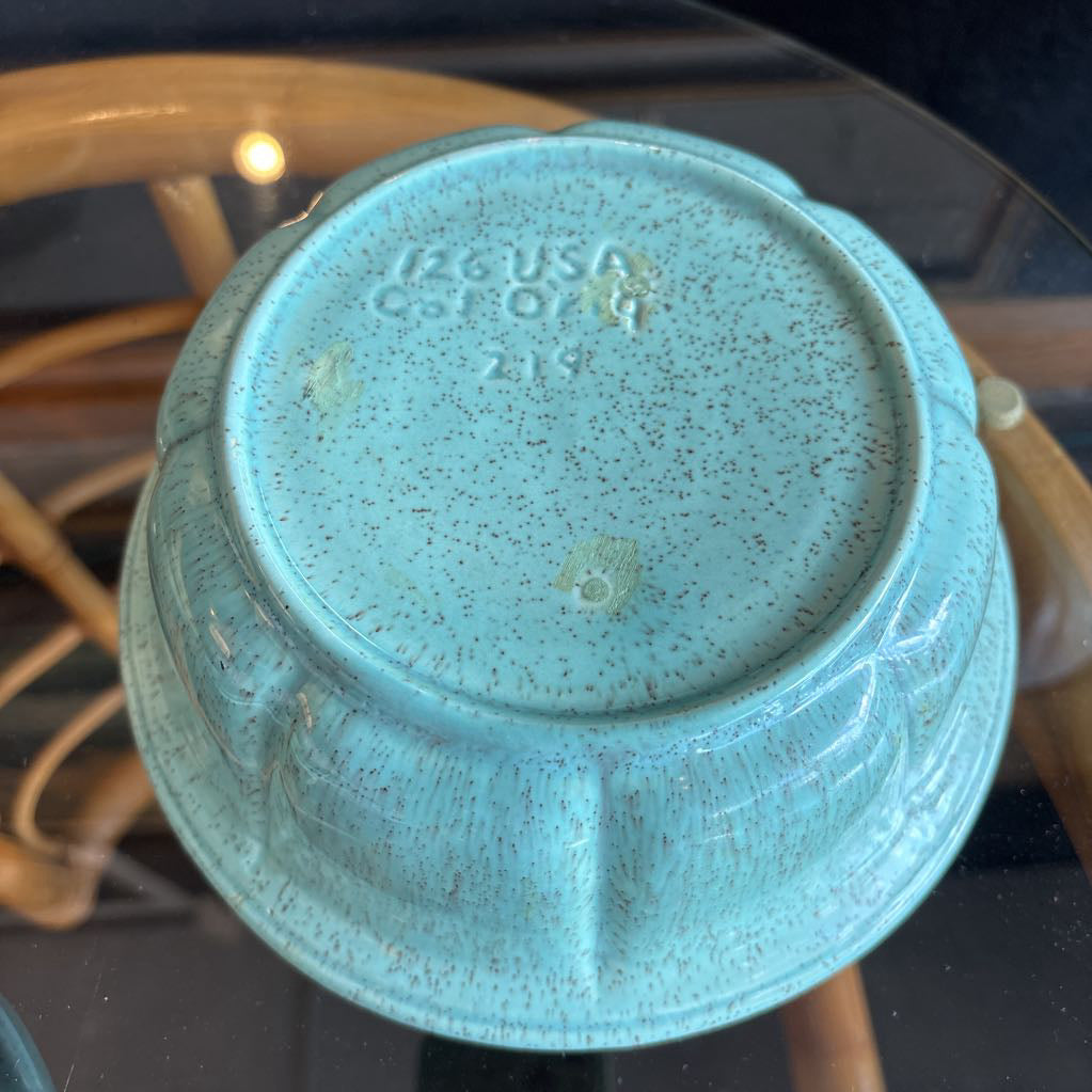 A ceramic turquoise serving bowl and lid in a brass wire caddy with handles on a glass table in front of a black wall.