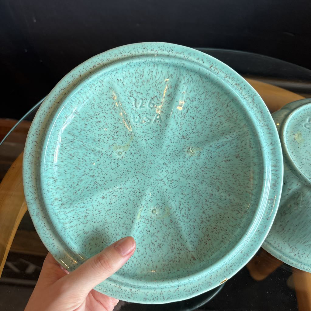 A ceramic turquoise serving bowl and lid in a brass wire caddy with handles on a glass table in front of a black wall.