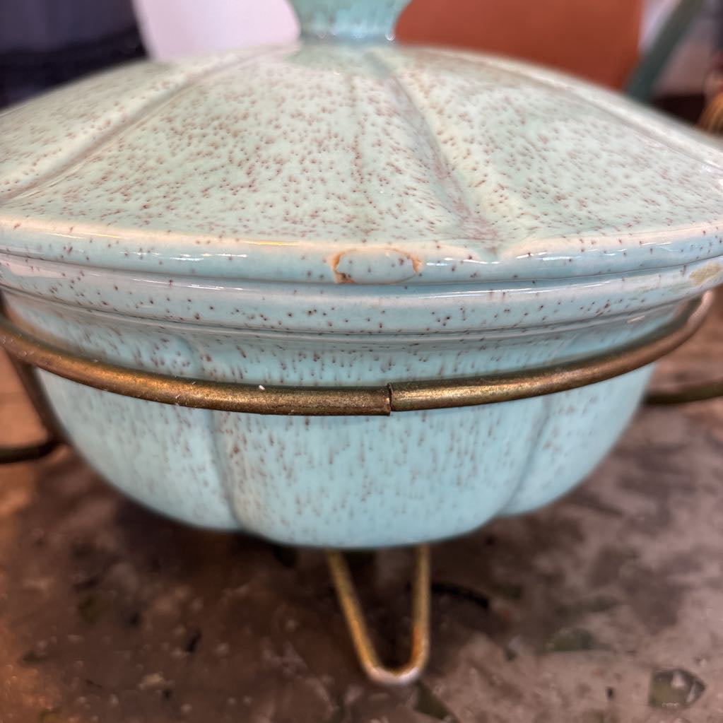 A ceramic turquoise serving bowl and lid in a brass wire caddy with handles on a glass table in front of a black wall.