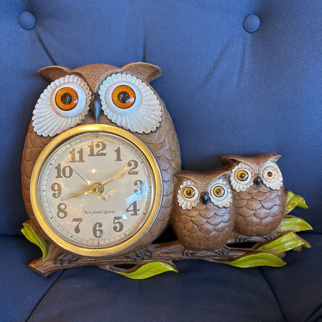A vintage clock of 3 owls with wide eyes sitting on a branch and the clock face in the largest owl. This clock is on a blue cushion. 