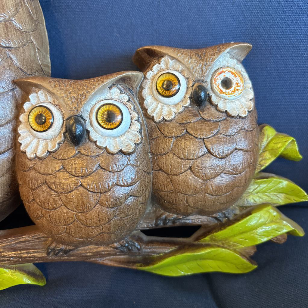 A vintage clock of 3 owls with wide eyes sitting on a branch and the clock face in the largest owl. This clock is on a blue cushion. 