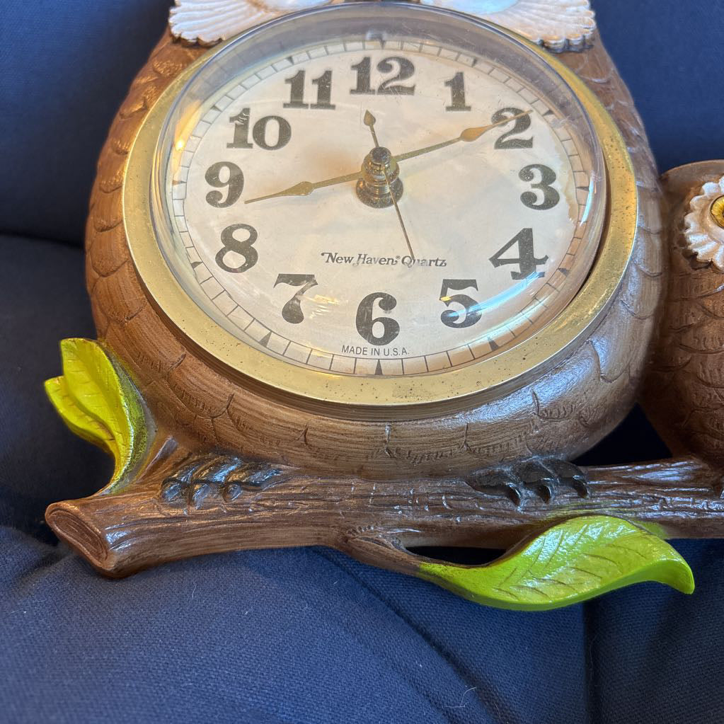 A vintage clock of 3 owls with wide eyes sitting on a branch and the clock face in the largest owl. This clock is on a blue cushion. 