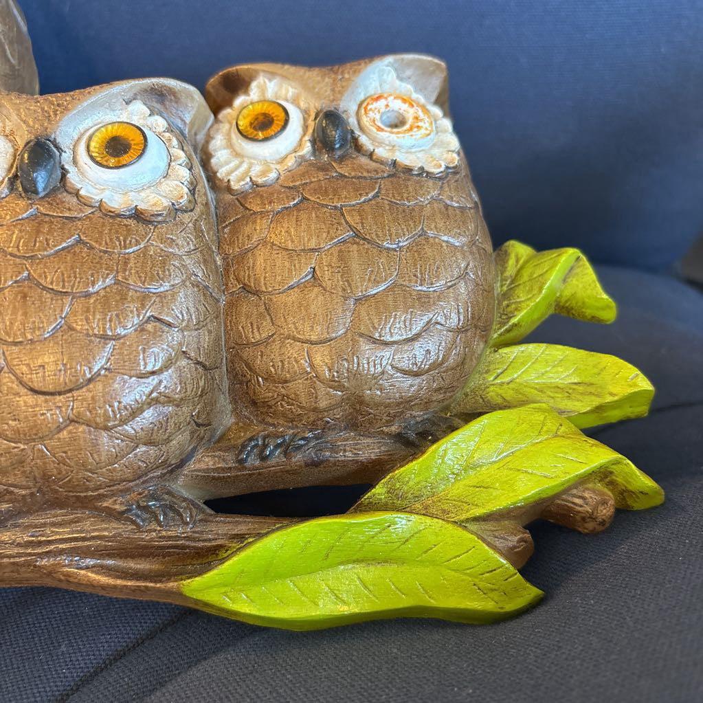 A vintage clock of 3 owls with wide eyes sitting on a branch and the clock face in the largest owl. This clock is on a blue cushion. 