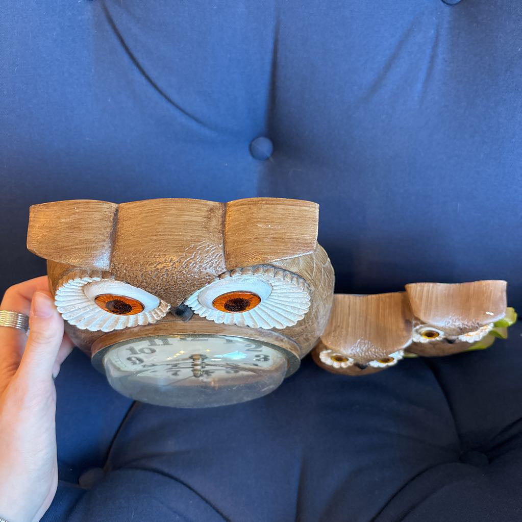 A vintage clock of 3 owls with wide eyes sitting on a branch and the clock face in the largest owl. This clock is on a blue cushion. 