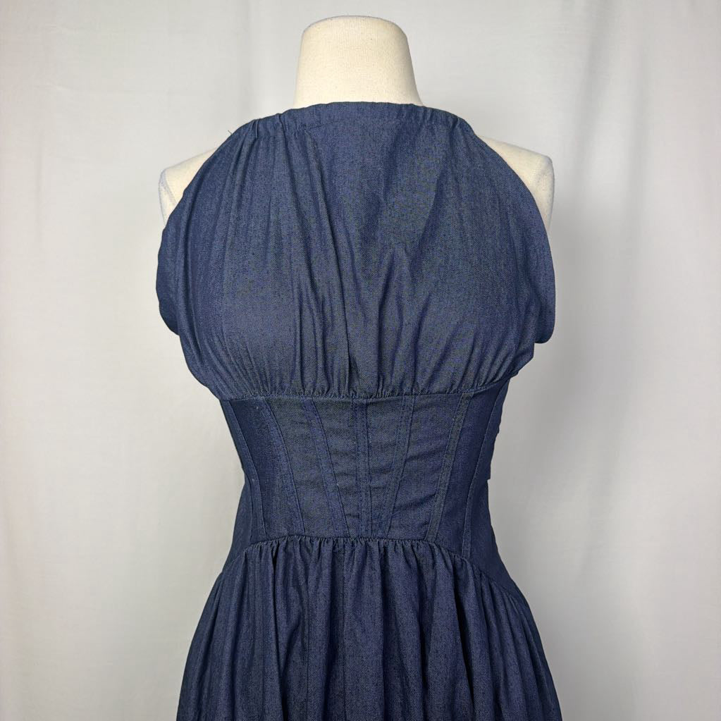 A sleeveless halter-neck dress in dark blue chambray fabric, shown from the back on a cream mannequin torso against a light gray fabric backdrop.