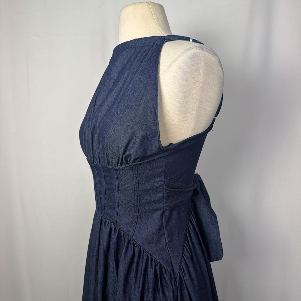 A sleeveless halter-neck dress in dark blue chambray fabric, shown from the back on a cream mannequin torso against a light gray fabric backdrop.