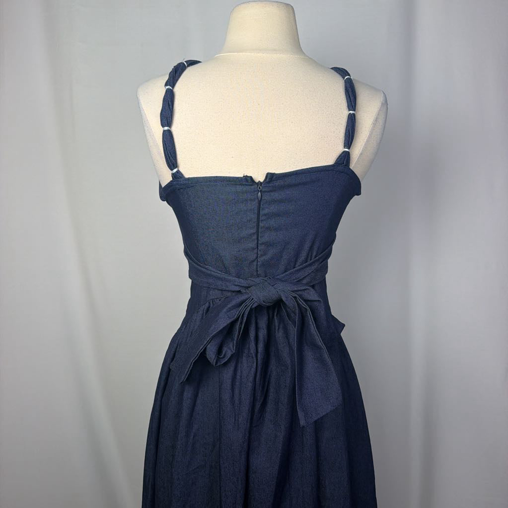 A navy blue denim dress displayed on a cream mannequin torso against a light gray fabric backdrop. The dress features thin shoulder straps with white accents, a square neckline, a gathered waist, and a self-tie belt at the back.