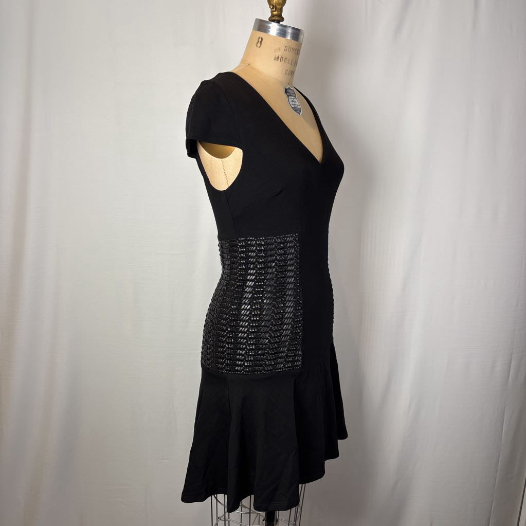 A black short-sleeve V-neck dress displayed on a dress form mannequin against a white fabric backdrop. The dress features a textured, patterned waist panel and a flared skirt.