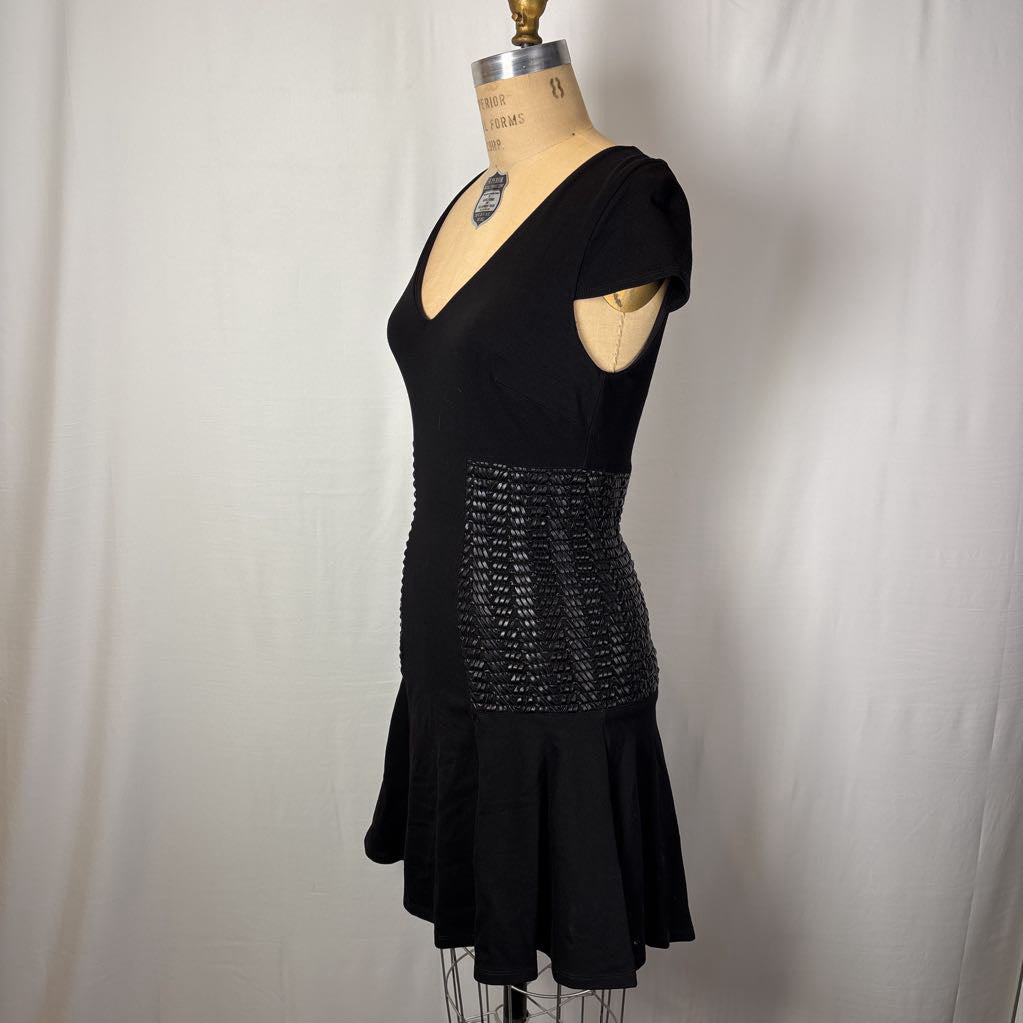 A black sleeveless dress displayed on a dress form mannequin against a white fabric backdrop. The dress has a V-neckline, short cap sleeves with cutouts, a textured beaded waist panel, and a flared skirt.