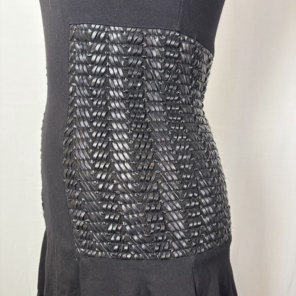 Close-up of a black dress or garment on a mannequin, showing a wide textured panel with a woven or braided chevron pattern across the torso.