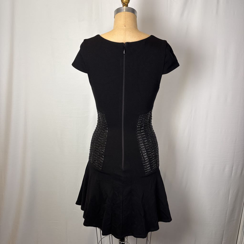A black short-sleeve fit-and-flare dress displayed on a dress form, showing a center zipper and textured side panels, against a white fabric backdrop.