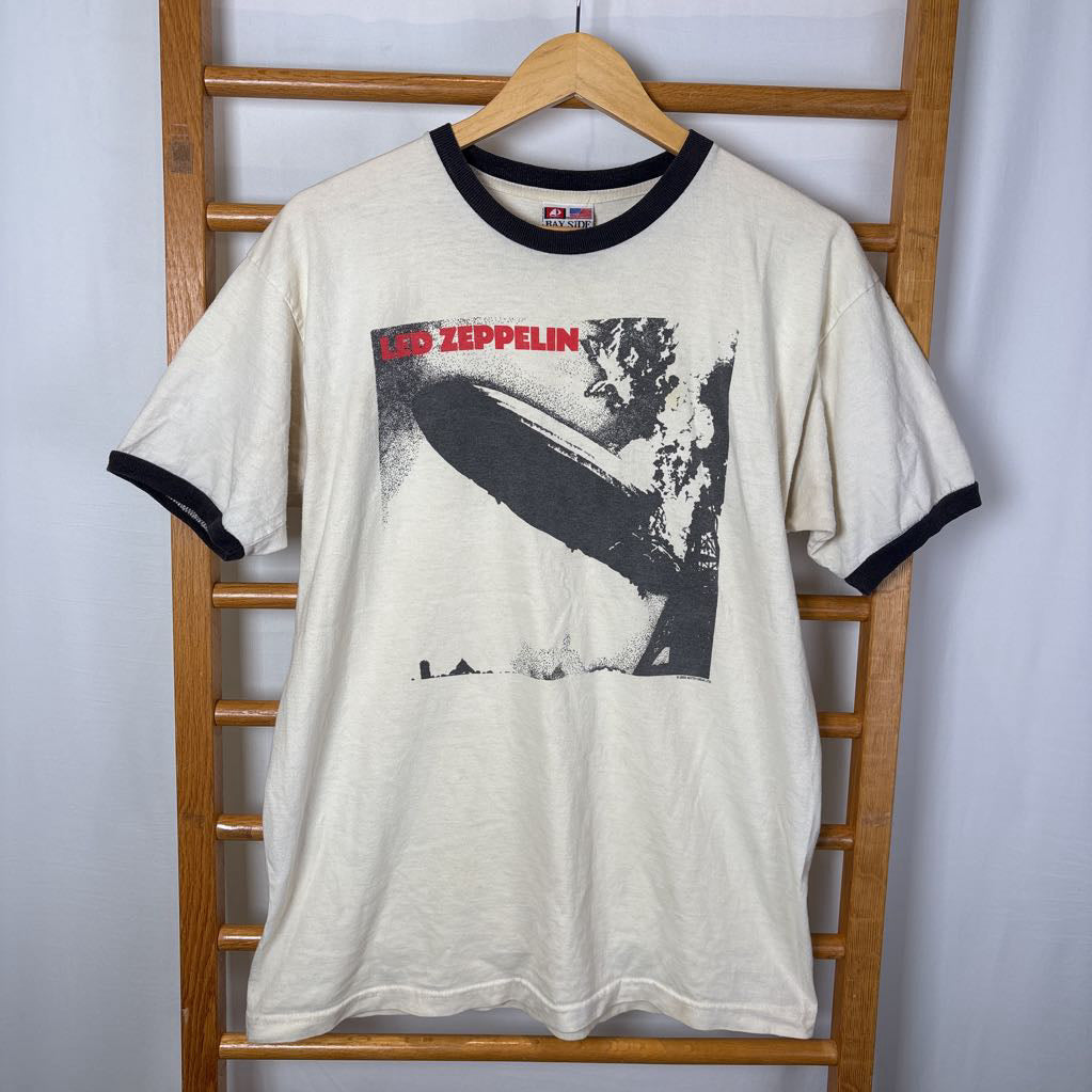 A white vintage style short sleeve t-shirt with an image of a Led Zeppelin album cover and the band's name in red letters. The shirt hangs on a wooden rack against a white background. 