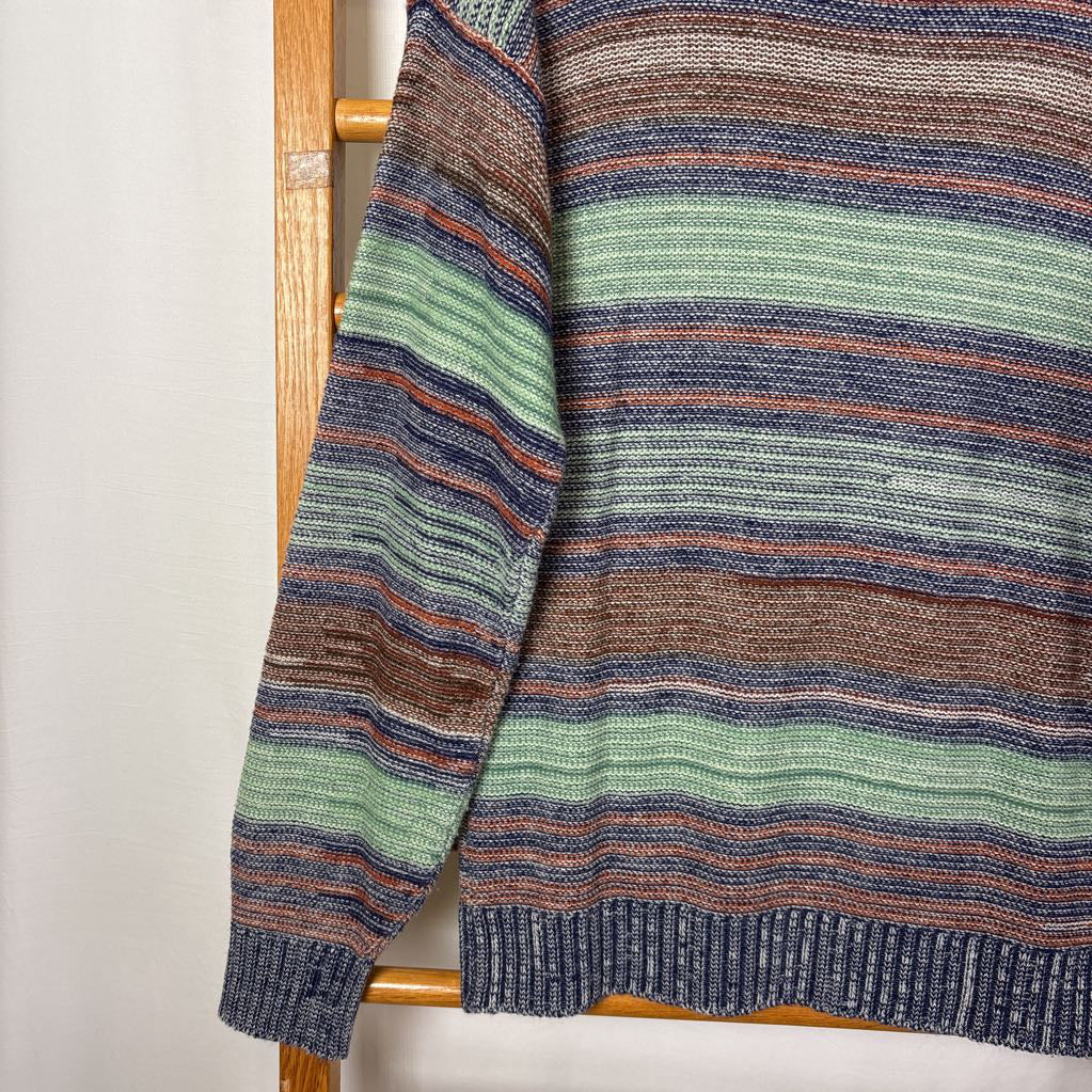 A blue, green, and brown striped knitted long sleeve sweater hanging on a wooden rack against a white background.