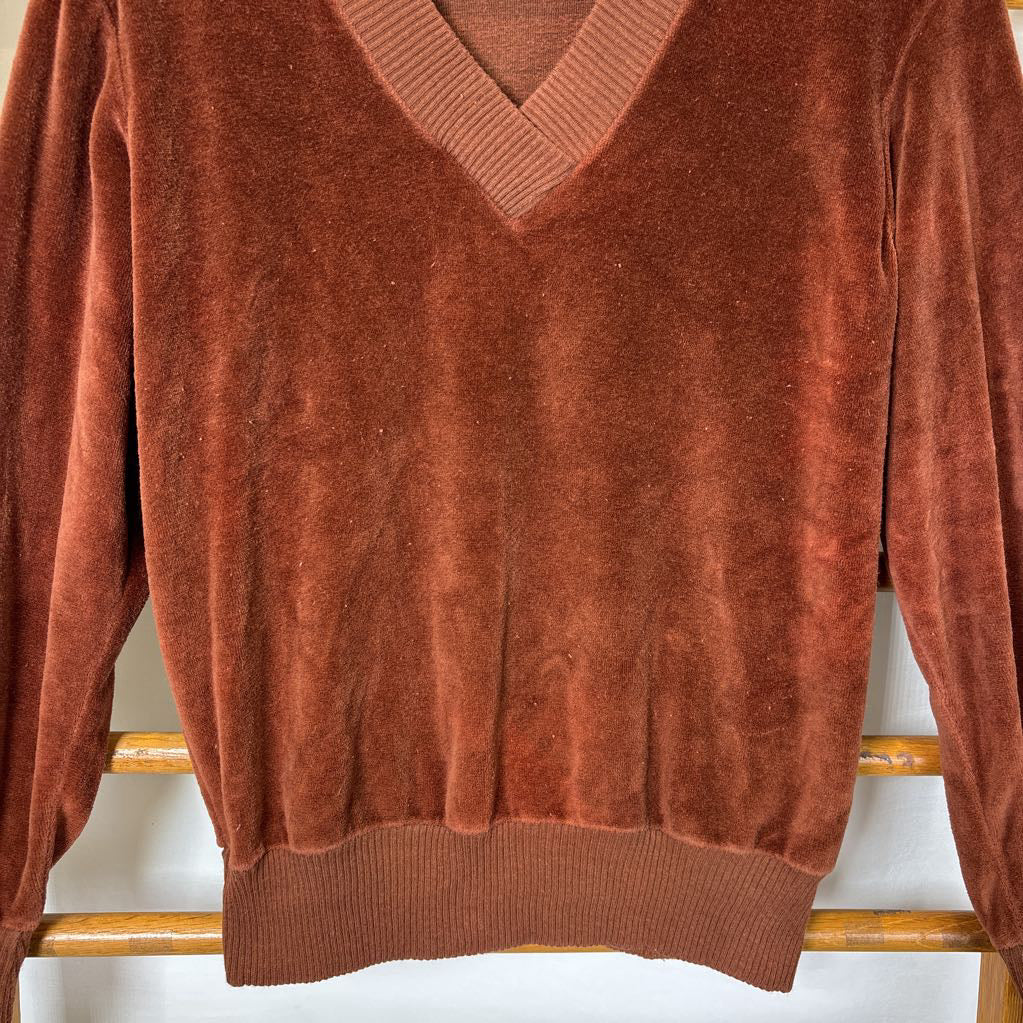 A burnt orange velour long sleeve V-neck sweater hanging on a wooden rack against a white wall.