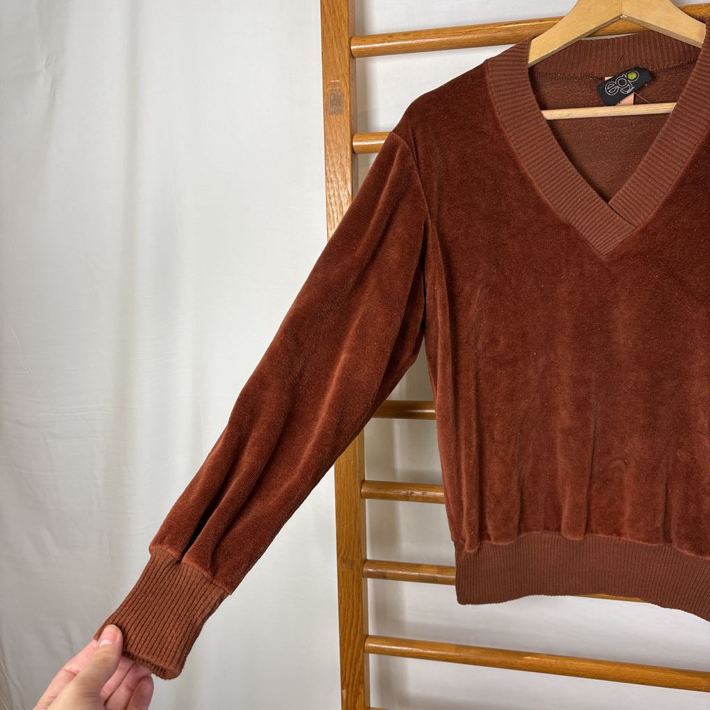 A burnt orange velour long sleeve V-neck sweater hanging on a wooden rack against a white wall.