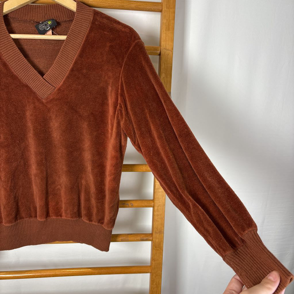 A burnt orange velour long sleeve V-neck sweater hanging on a wooden rack against a white wall.