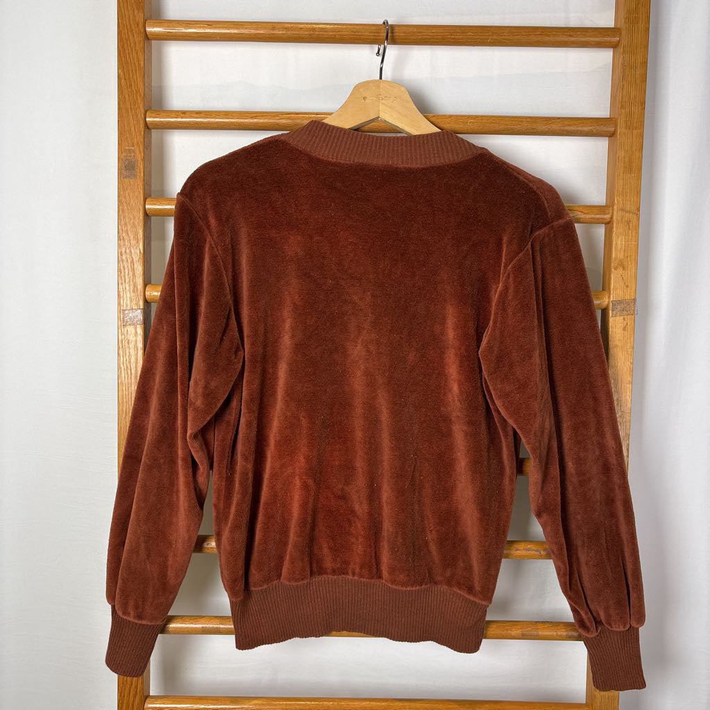 A burnt orange velour long sleeve V-neck sweater hanging on a wooden rack against a white wall.