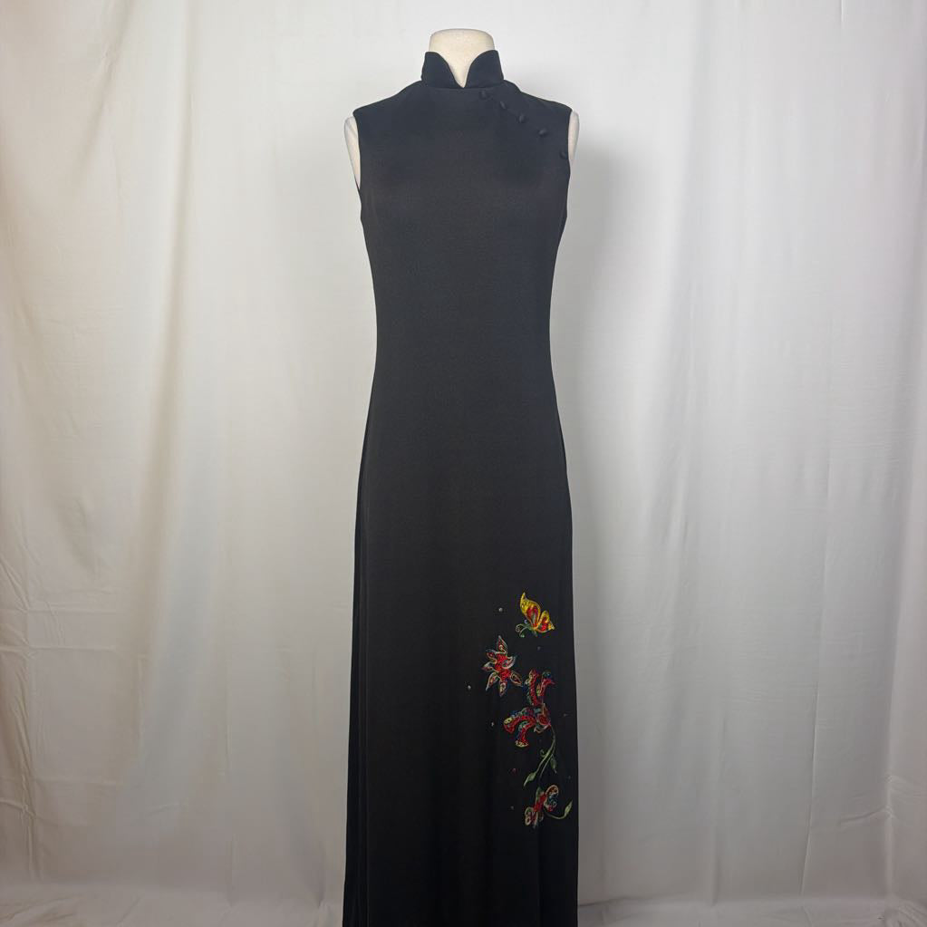 A black maxi dress with a butterfly and floral embroidery at the bottom. This dress is on a mannequin in front of a white wall. 