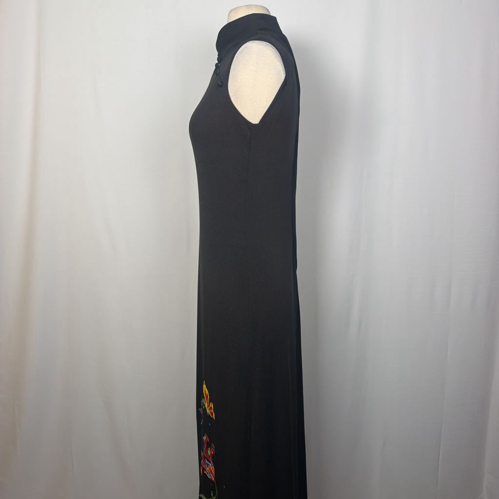 A sleeveless black maxi dress with a mock neck and side cutouts, displayed on a mannequin against a white fabric backdrop. The dress features colorful embroidered floral details near the lower hem.