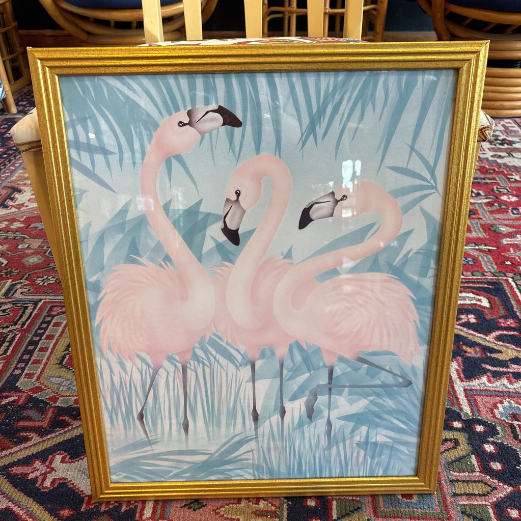 A print of three pink flamingos in a tropical setting in a gold frame in a room setting.
