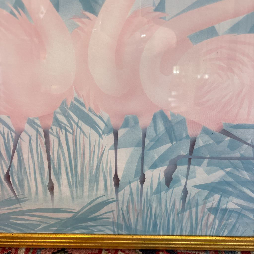 A print of three pink flamingos in a tropical setting in a gold frame in a room setting.