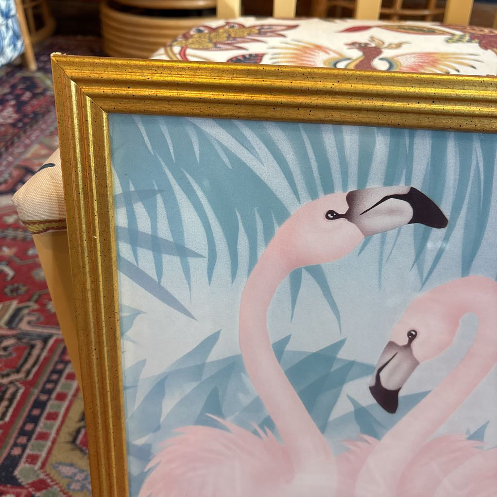 A print of three pink flamingos in a tropical setting in a gold frame in a room setting.