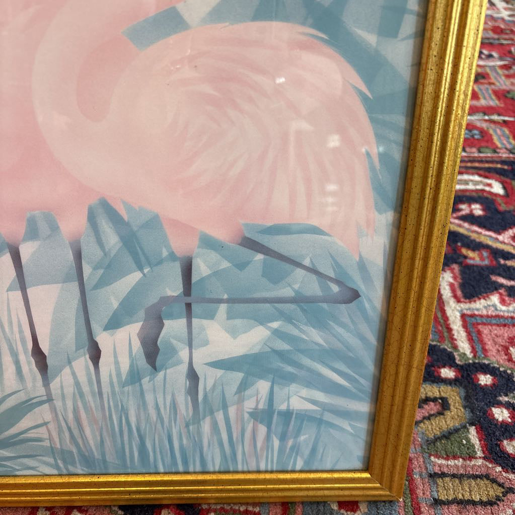 A print of three pink flamingos in a tropical setting in a gold frame in a room setting.