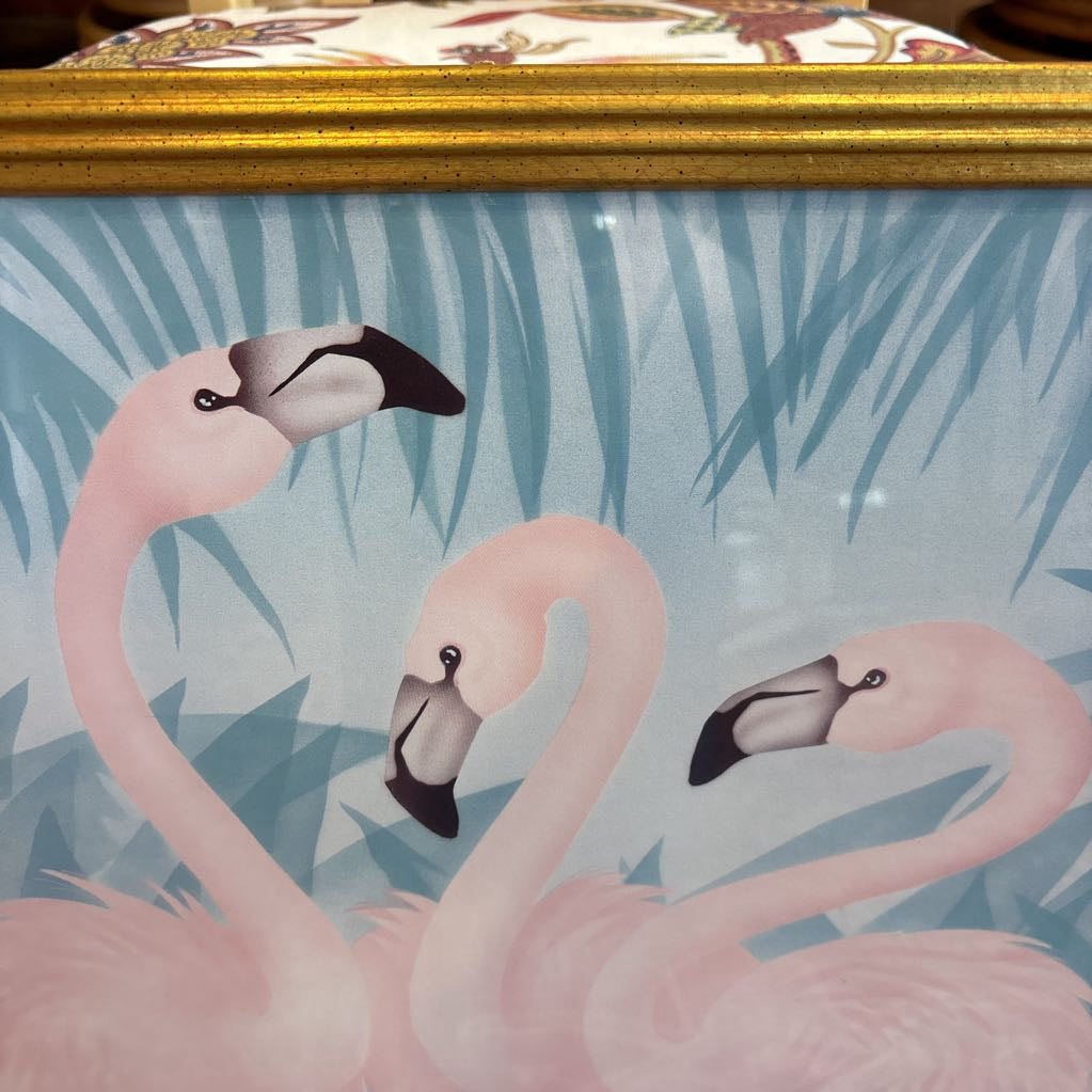 A print of three pink flamingos in a tropical setting in a gold frame in a room setting.