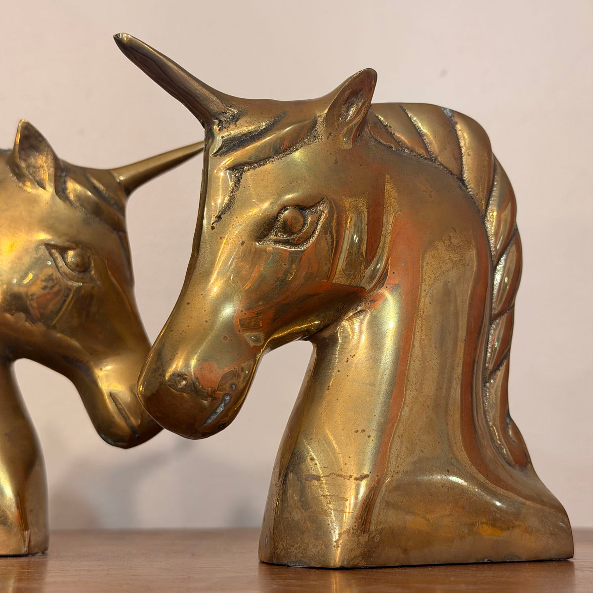 A close-up of a metallic gold unicorn head sculpture on a wooden surface against a plain light background, with part of a second similar unicorn visible on the left.