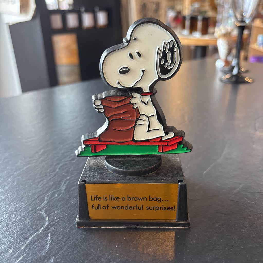 A small figure of snoopy on a pedestal. He's opening up a brown bag and the phrase at the bottom reads, "Life is like a brown bag...full of wonderful surprises!" 