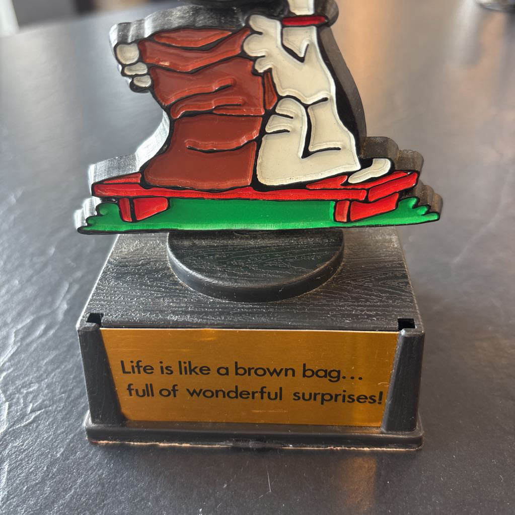 A small figure of snoopy on a pedestal. He's opening up a brown bag and the phrase at the bottom reads, "Life is like a brown bag...full of wonderful surprises!" 