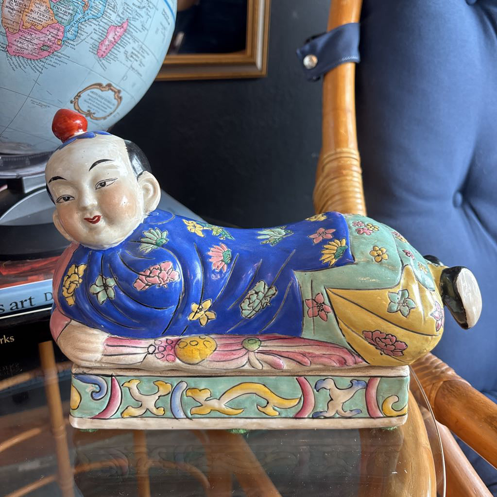 A hand-painted figure of a reclining boy dressed in a vivid royal blue robe adorned with delicate floral motifs and a decorative pedestal base.