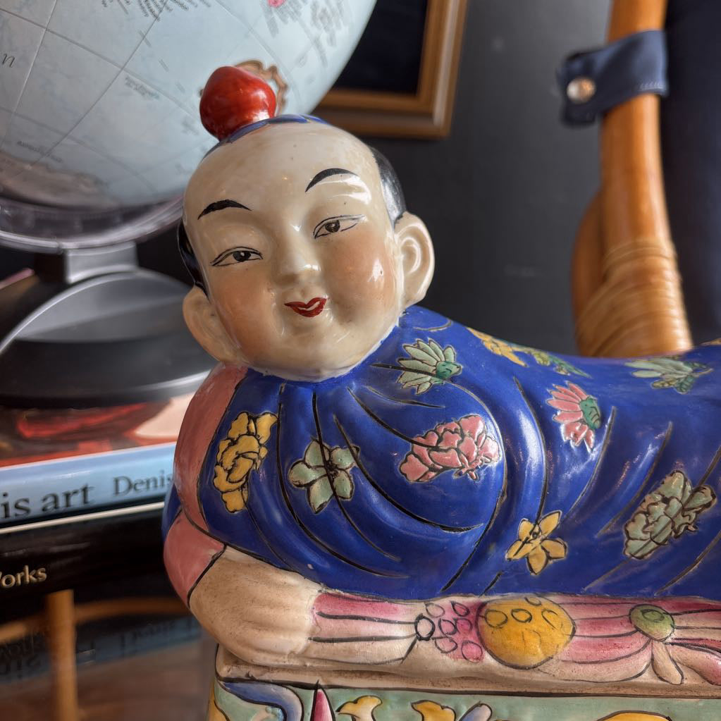 A hand-painted figure of a reclining boy dressed in a vivid royal blue robe adorned with delicate floral motifs and a decorative pedestal base.