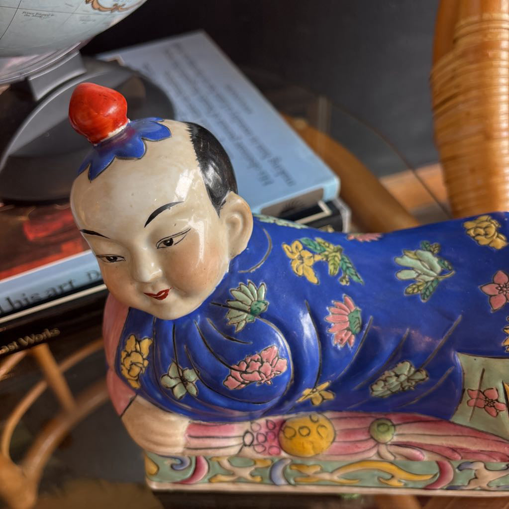 A hand-painted figure of a reclining boy dressed in a vivid royal blue robe adorned with delicate floral motifs and a decorative pedestal base.