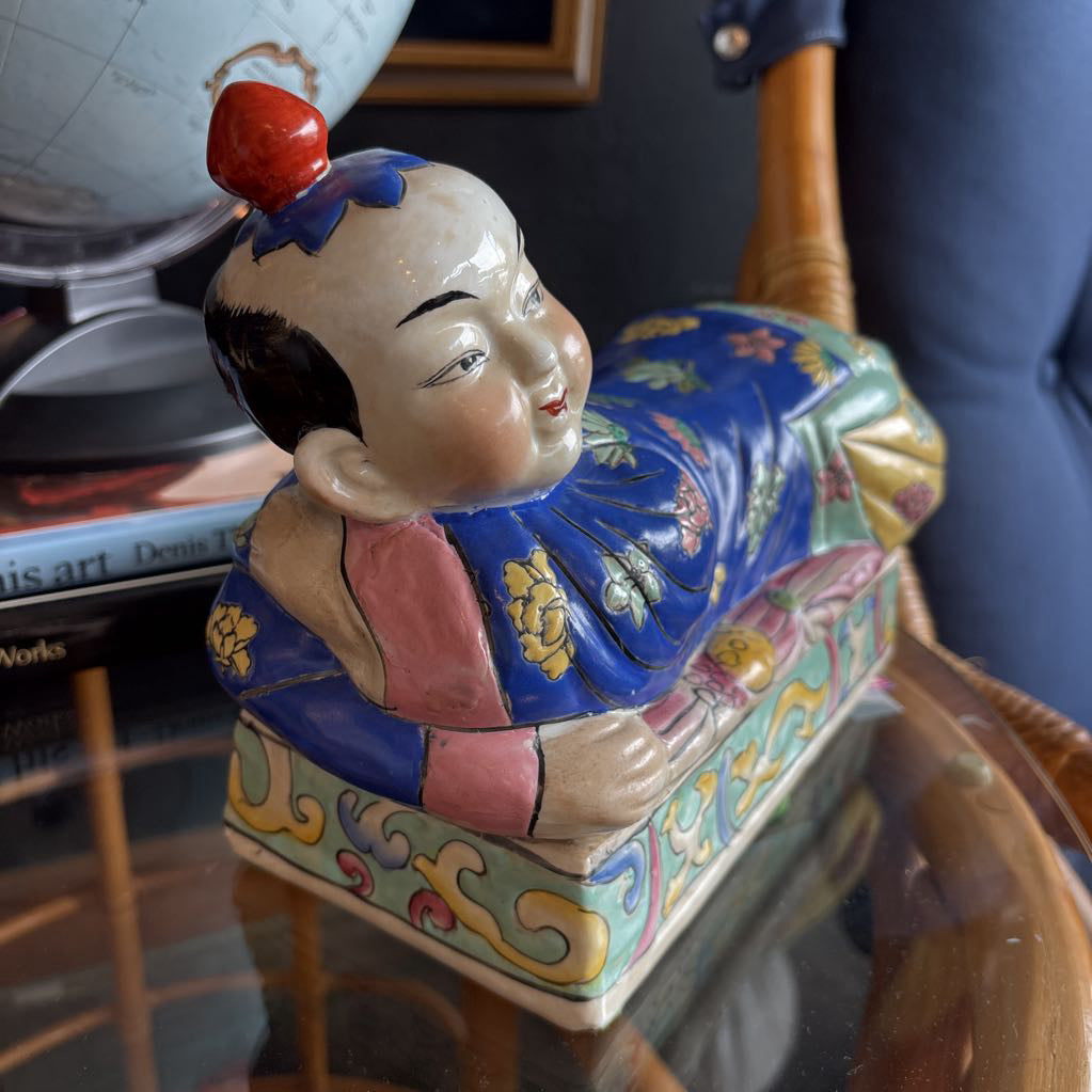 A hand-painted figure of a reclining boy dressed in a vivid royal blue robe adorned with delicate floral motifs and a decorative pedestal base.