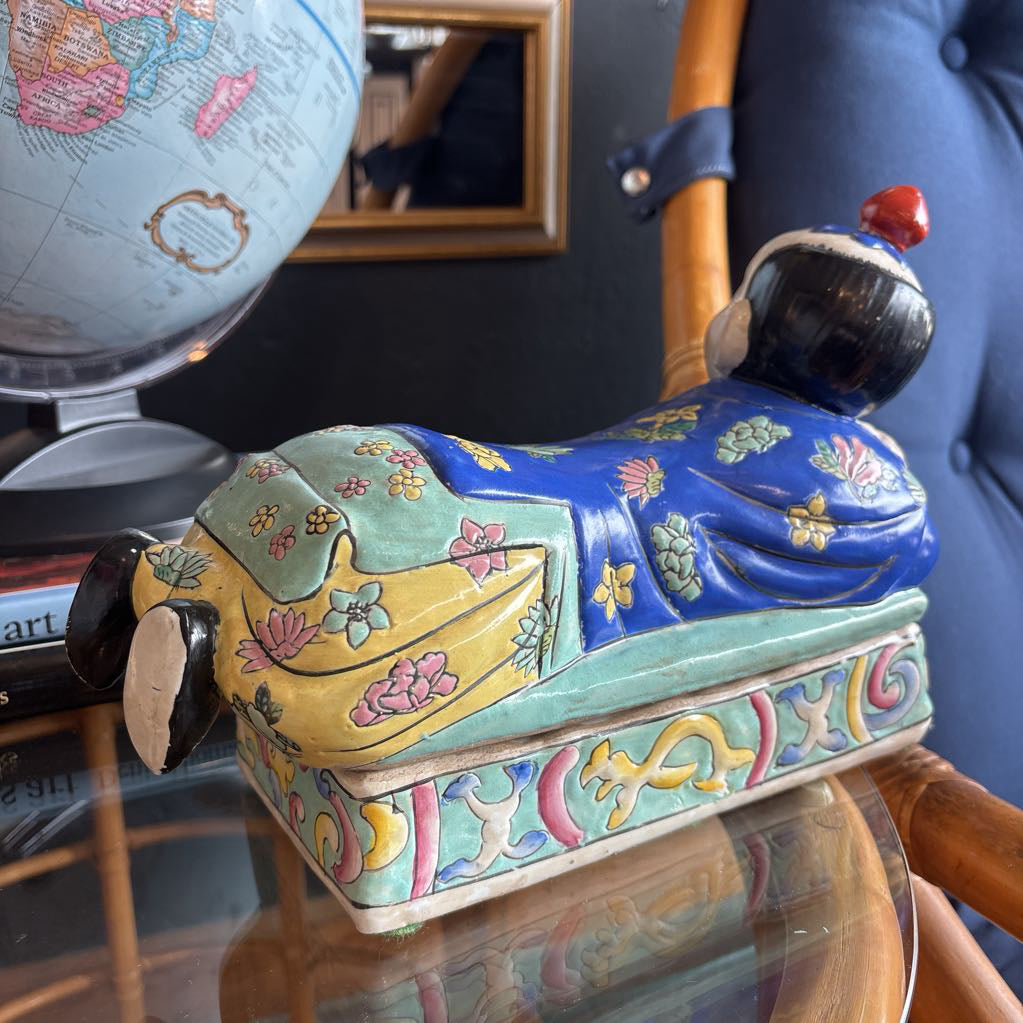 A hand-painted figure of a reclining boy dressed in a vivid royal blue robe adorned with delicate floral motifs and a decorative pedestal base.