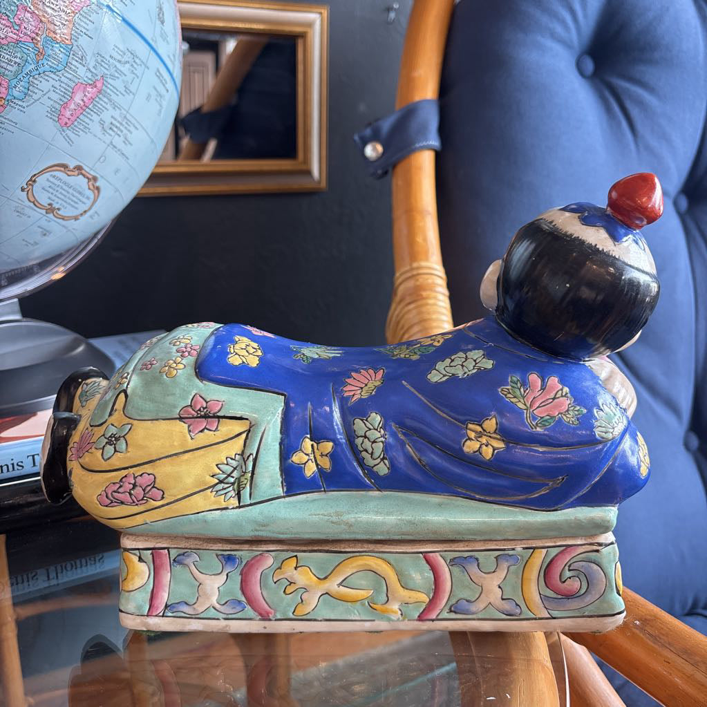 A hand-painted figure of a reclining boy dressed in a vivid royal blue robe adorned with delicate floral motifs and a decorative pedestal base.