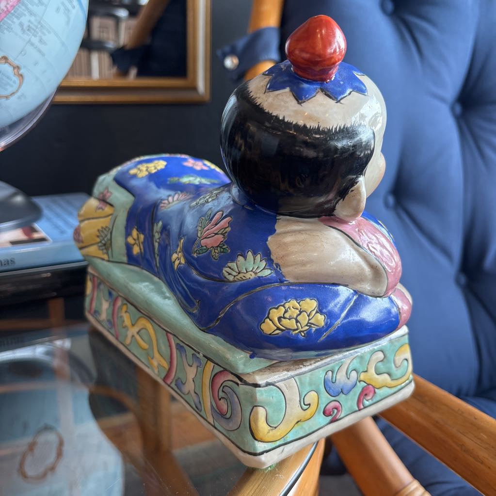 A hand-painted figure of a reclining boy dressed in a vivid royal blue robe adorned with delicate floral motifs and a decorative pedestal base.