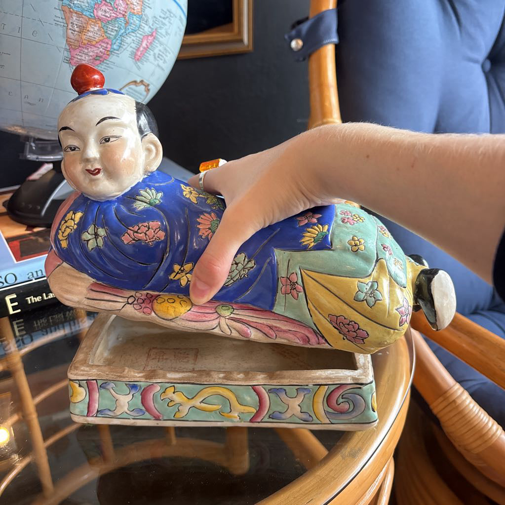A hand-painted figure of a reclining boy dressed in a vivid royal blue robe adorned with delicate floral motifs and a decorative pedestal base being opened by an arm to show its a container.
