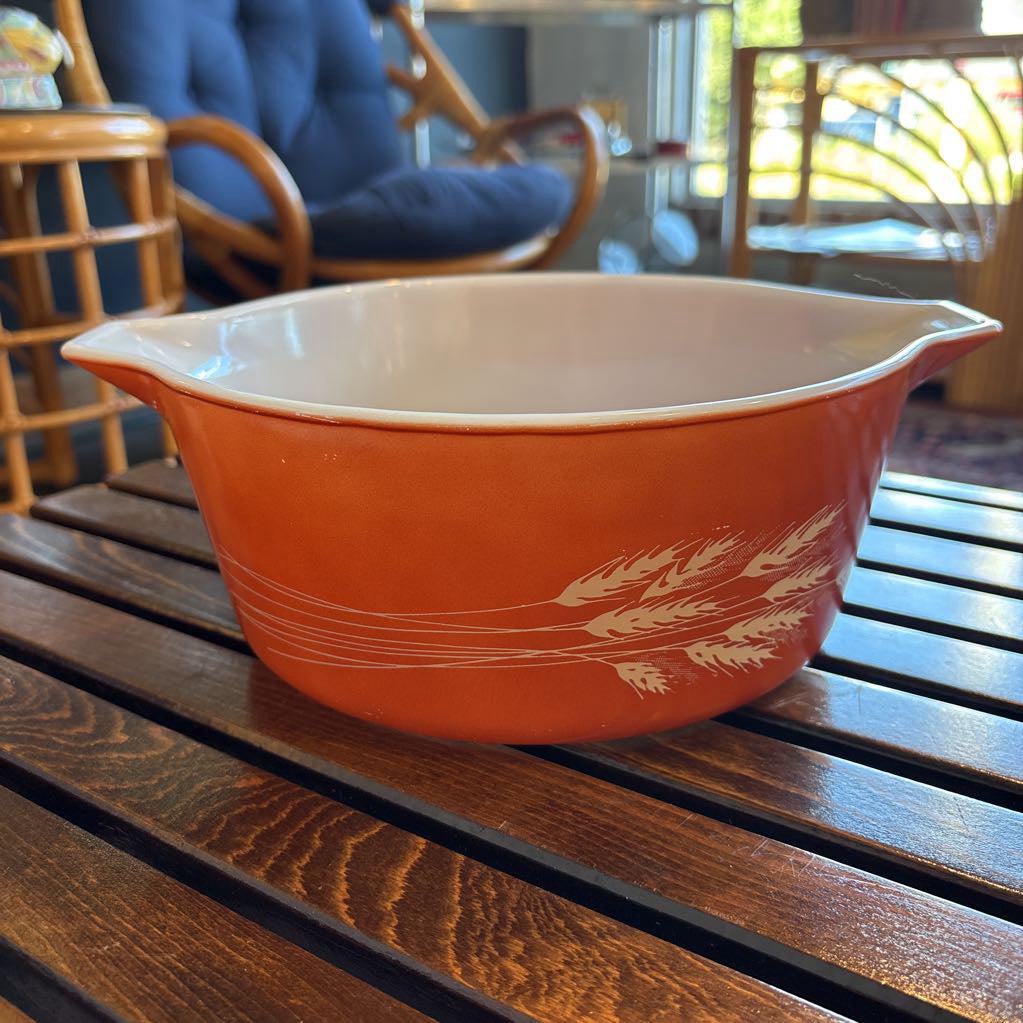 An orange pyrex casserole dish with the imagery of stalks of wheat across the exterior. The dish sits on a wooden slat bench in a room setting. 