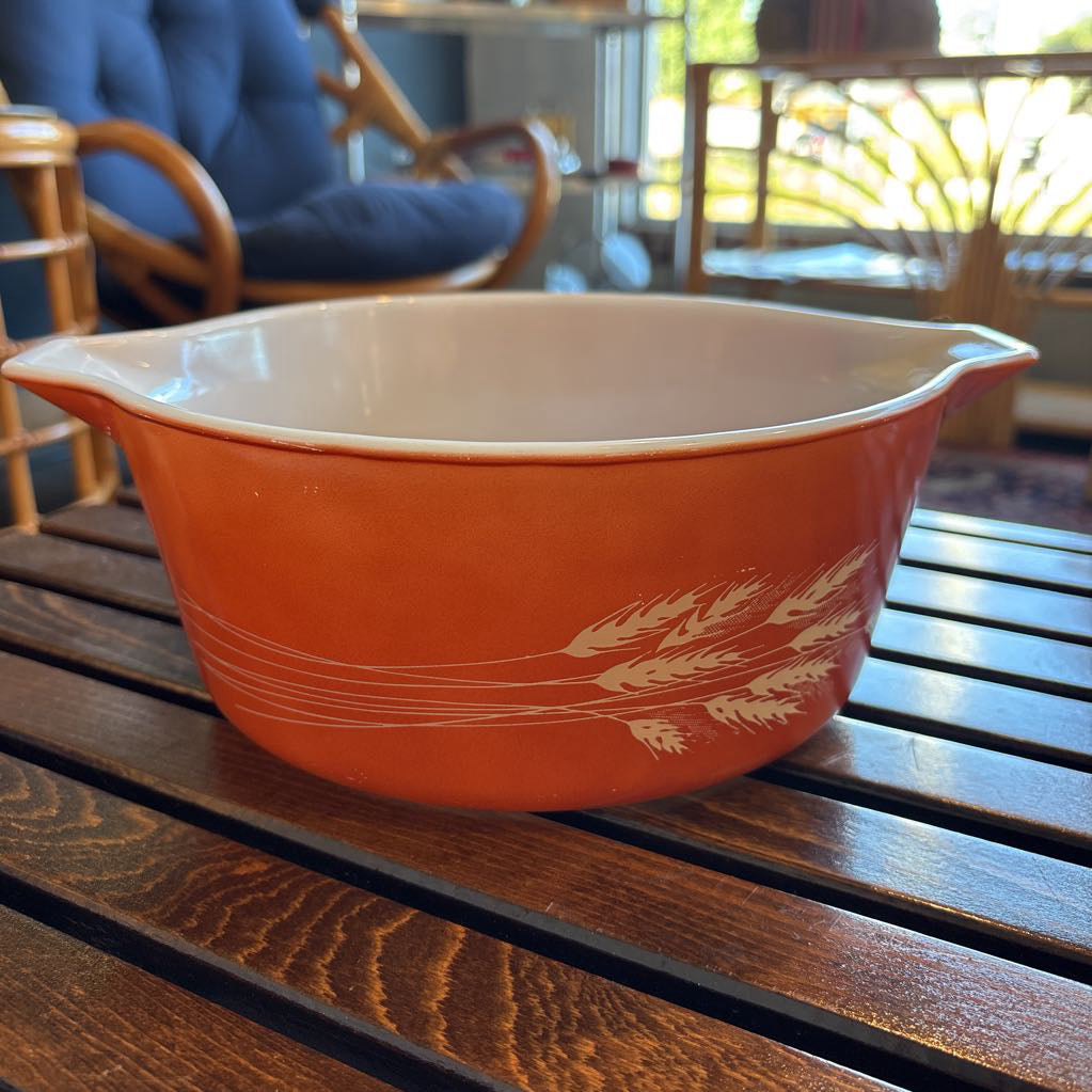 An orange pyrex casserole dish with the imagery of stalks of wheat across the exterior. The dish sits on a wooden slat bench in a room setting. 