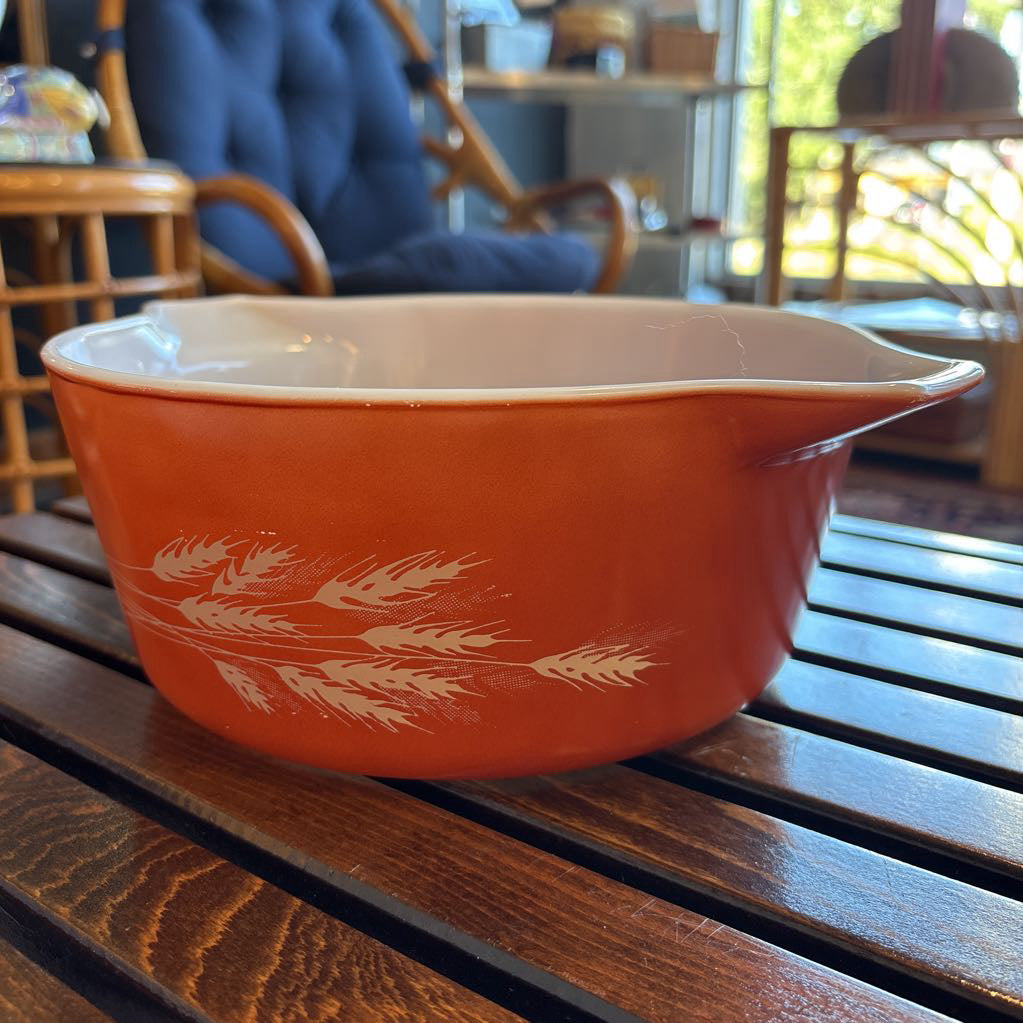 An orange pyrex casserole dish with the imagery of stalks of wheat across the exterior. The dish sits on a wooden slat bench in a room setting. 
