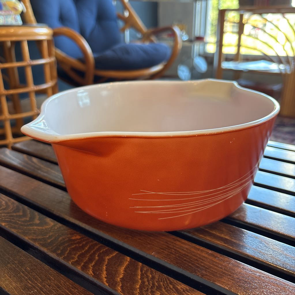 An orange pyrex casserole dish with the imagery of stalks of wheat across the exterior. The dish sits on a wooden slat bench in a room setting. 