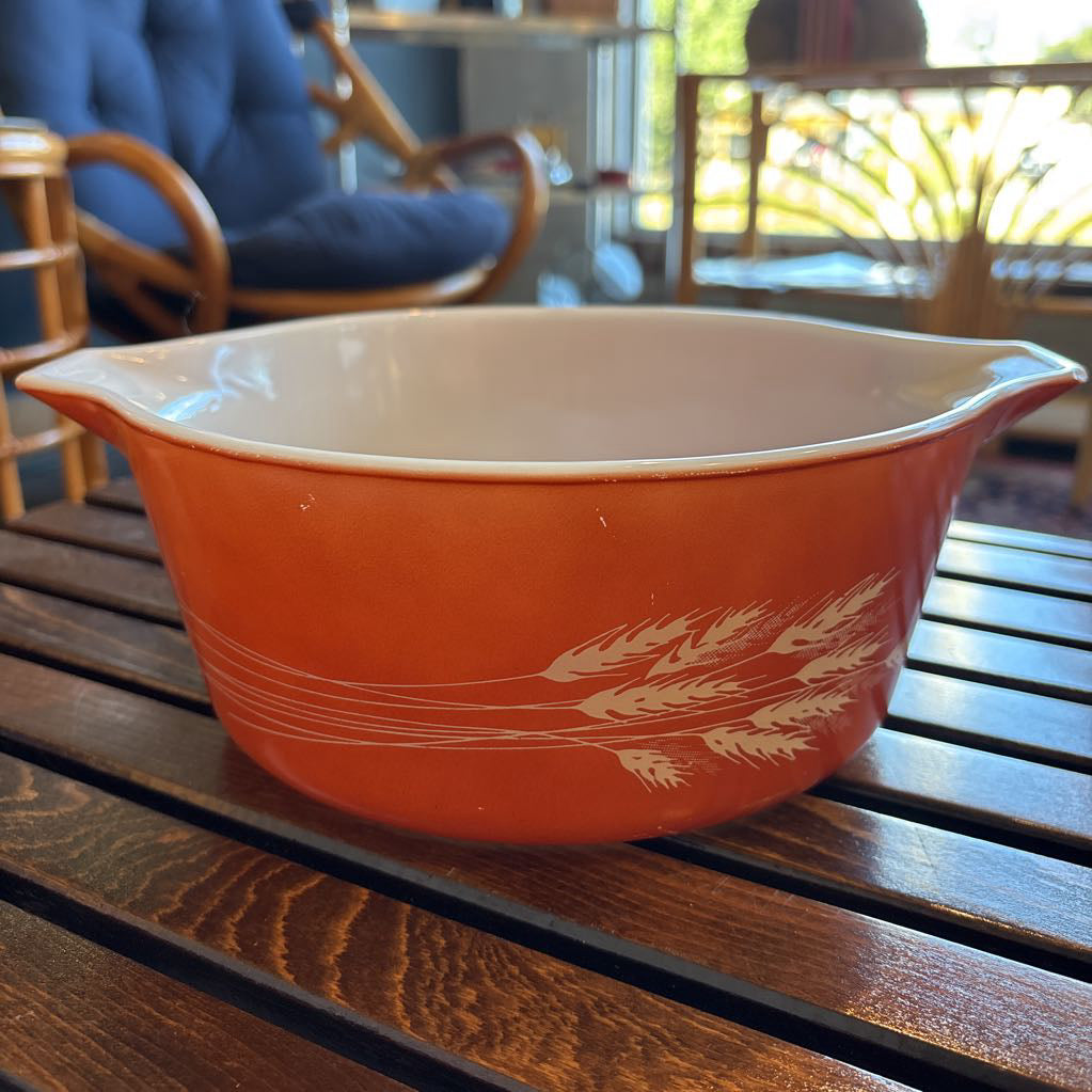 An orange pyrex casserole dish with the imagery of stalks of wheat across the exterior. The dish sits on a wooden slat bench in a room setting. 