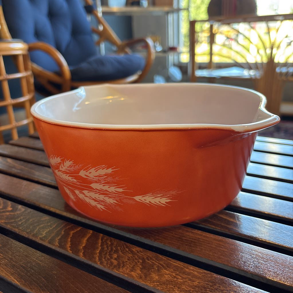 An orange pyrex casserole dish with the imagery of stalks of wheat across the exterior. The dish sits on a wooden slat bench in a room setting. 