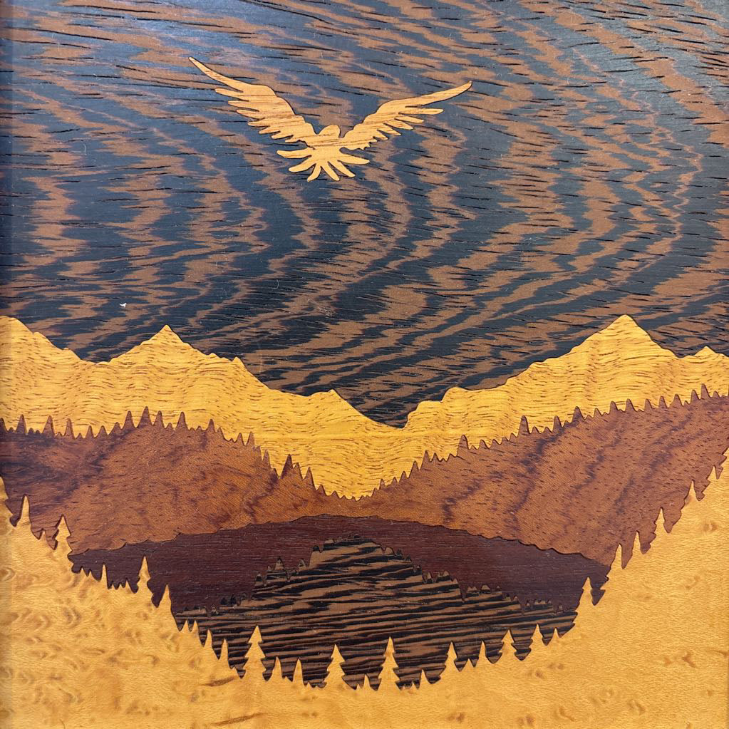 A square framed wooden inlaid artwork of a bird flying over mountains and forests with different colors and textures of wood. 