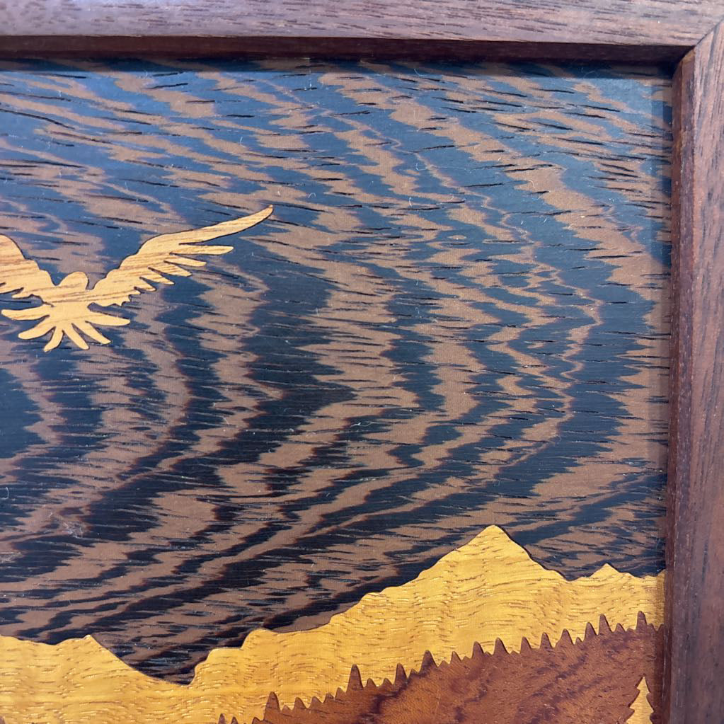 A square framed wooden inlaid artwork of a bird flying over mountains and forests with different colors and textures of wood. 
