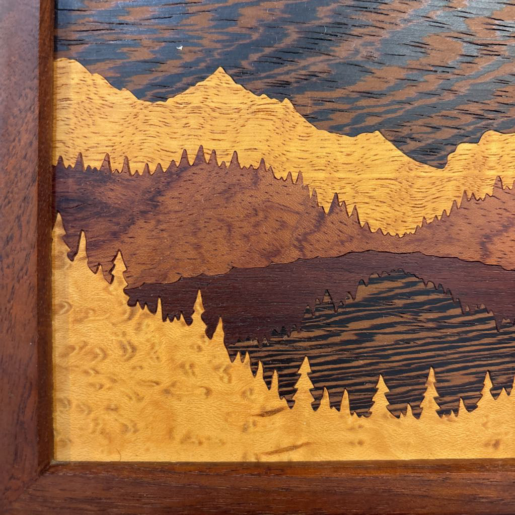 A square framed wooden inlaid artwork of a bird flying over mountains and forests with different colors and textures of wood. 